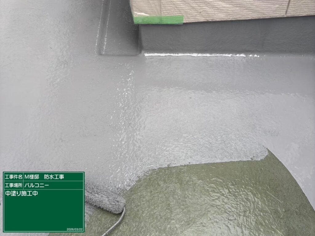 【FRP防水 トップコート1回目 施工中】<br />
1回目となる高伸縮・高耐候性トップコートの塗布作業です。ベランダ床面は直射日光や雨水、歩行による摩擦を最も受ける過酷な場所のため、膜厚が不均一にならないよう一定のペースで丁寧に塗り進めます。